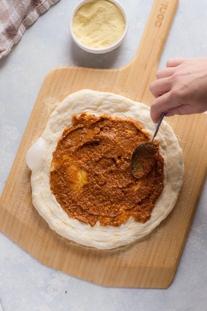 Pizza dough on a peel being spread with red sauce using a spoon. A small bowl of cornmeal is placed above.