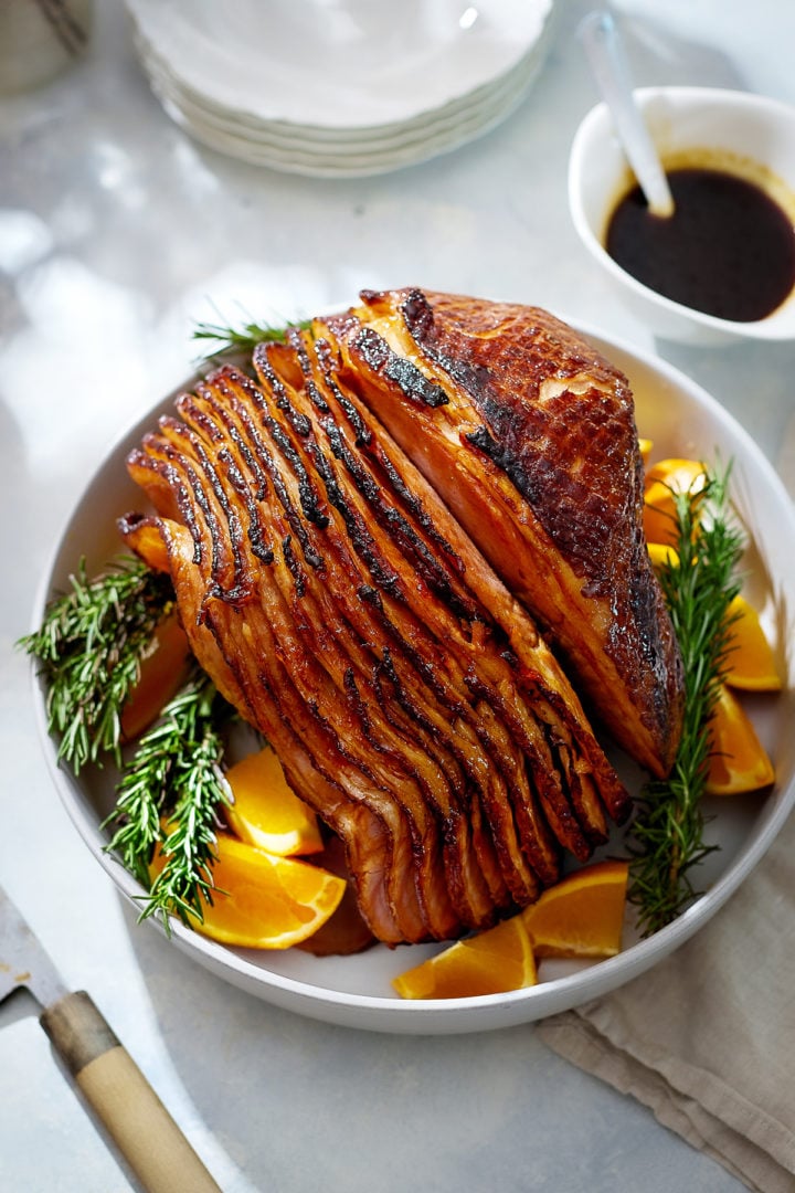 A close-up view of a fully cooked orange glazed ham resting on a white serving platter, surrounded by rosemary sprigs and orange wedges. The glaze gives the ham a shiny, amber-brown finish with slightly crisped edges.

