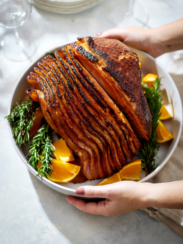 Hands are holding a white serving platter with a spiral-cut orange glazed ham in the center. The ham is coated in a glossy brown sugar spice glaze and surrounded by fresh rosemary sprigs and wedges of bright orange.