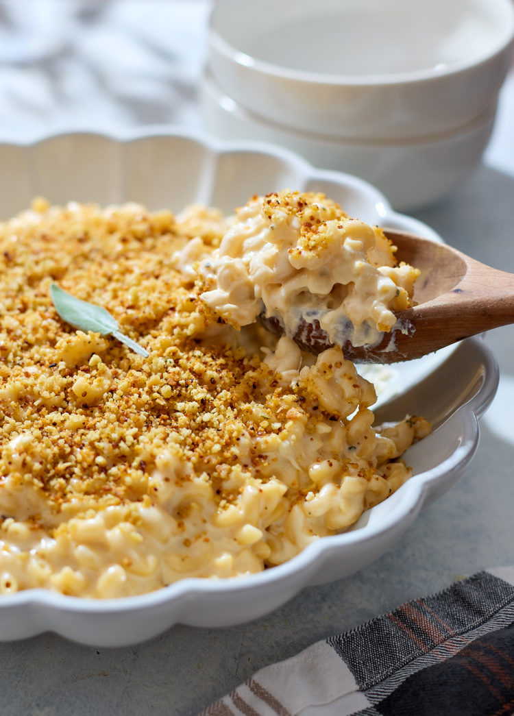 A close-up of three cheese mac and cheese in a white scalloped baking dish. A wooden spoon lifts a serving, showing creamy noodles coated in a thick cheese sauce beneath a golden breadcrumb topping. A single sage leaf sits on top of the crust, and soft natural light highlights the contrast between the crispy topping and the creamy interior.
