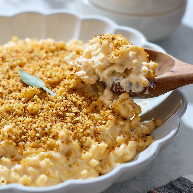 A close-up of three cheese mac and cheese in a white scalloped baking dish. A wooden spoon lifts a serving, showing creamy noodles coated in a thick cheese sauce beneath a golden breadcrumb topping. A single sage leaf sits on top of the crust, and soft natural light highlights the contrast between the crispy topping and the creamy interior.