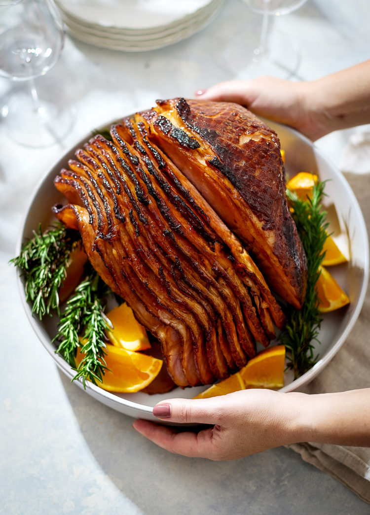 Hands are holding a white serving platter with a spiral-cut orange glazed ham in the center. The ham is coated in a glossy brown sugar spice glaze and surrounded by fresh rosemary sprigs and wedges of bright orange.
