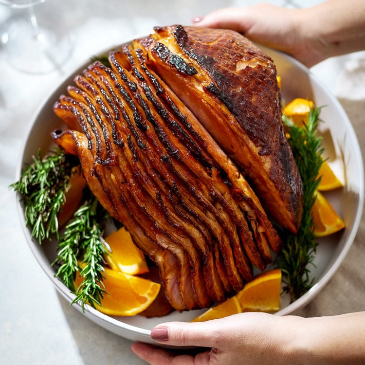 Hands are holding a white serving platter with a spiral-cut orange glazed ham in the center. The ham is coated in a glossy brown sugar spice glaze and surrounded by fresh rosemary sprigs and wedges of bright orange.