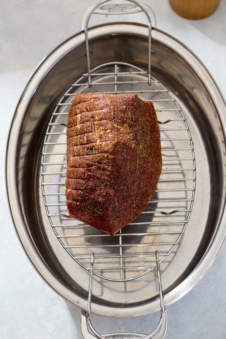 The seasoned beef roast, coated in a dark spice rub, rests on a metal roasting rack inside a stainless-steel roasting pan. The roast’s surface appears dry and evenly covered, ready for slow roasting.