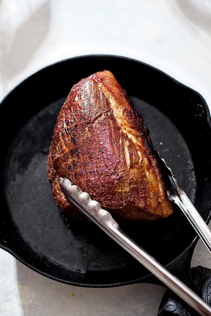 A browned eye of round roast sits in a cast iron skillet, being turned with metal tongs. The meat’s crust is caramelized and golden brown from searing after roasting, showing a rich texture under bright natural light.
