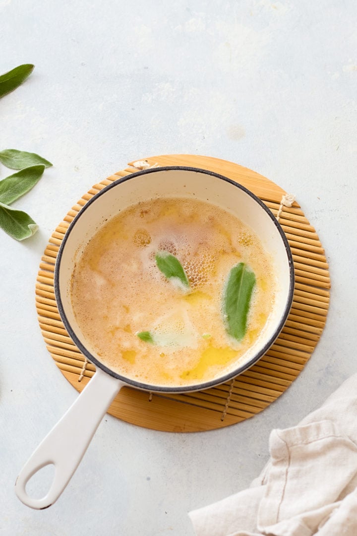 A small white saucepan with browned butter and floating sage leaves rests on a bamboo trivet. The melted butter has a light golden hue with a few foamy bubbles on top. Loose sage leaves and a beige linen napkin surround the pan on a light surface.