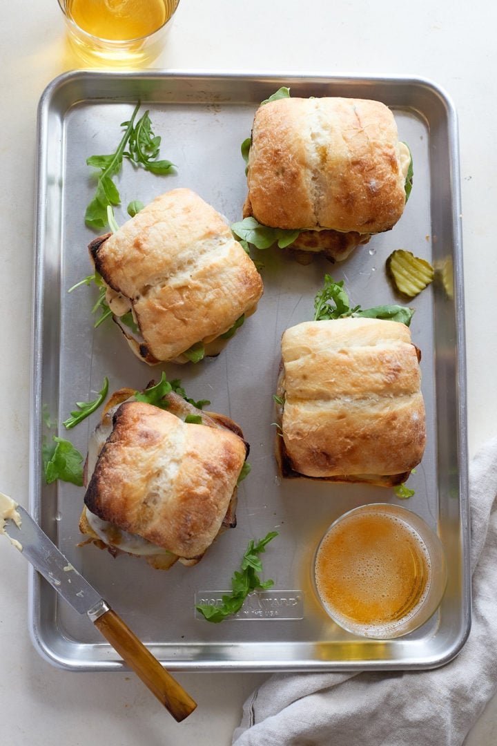 Four toasted hot roast beef sandwiches sit on a metal sheet pan. Each ciabatta roll is filled with roast beef, melted provolone, arugula, and pickles. A drink and a butter knife rest nearby, along with scattered arugula leaves.
