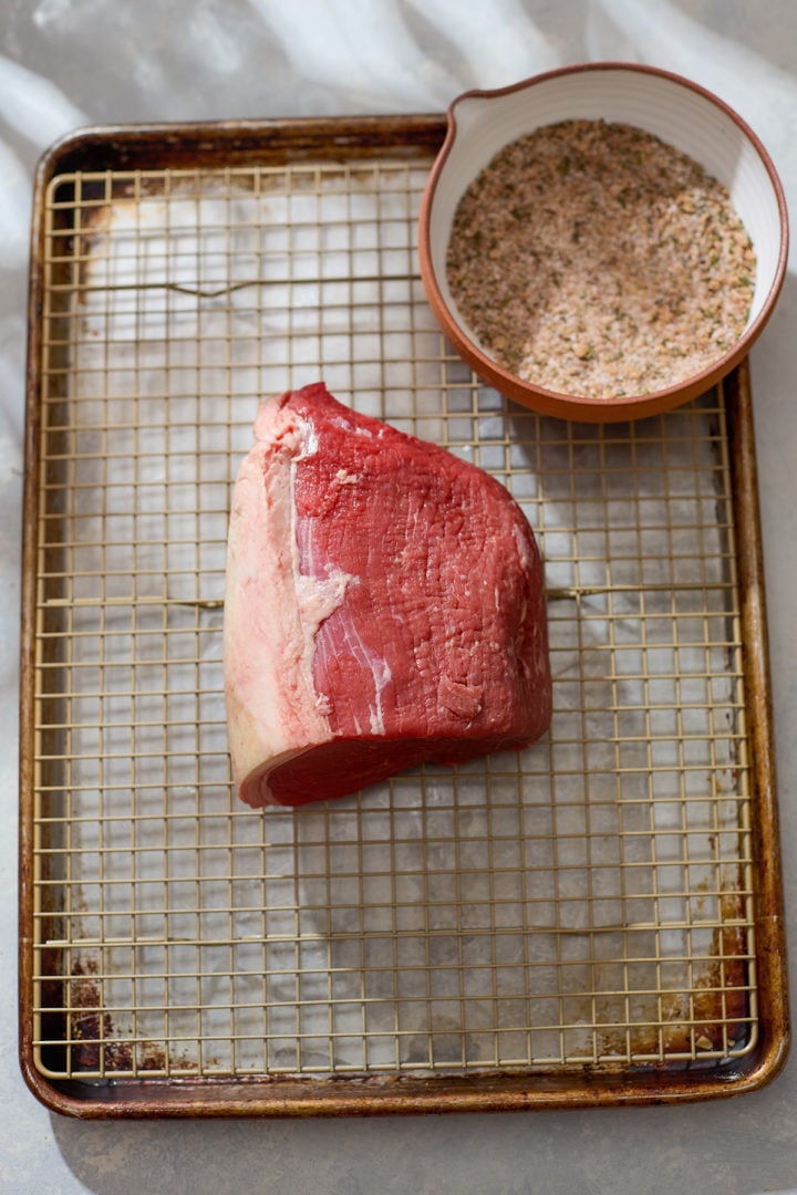 A raw eye of round roast sits on a wire rack over a baking sheet. Next to it is a small bowl filled with the blended seasoning mixture, ready to be rubbed over the meat. The scene is softly lit on a neutral surface.