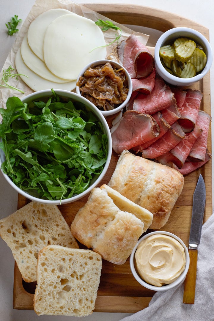 A wooden board is arranged with ingredients for homemade roast beef sandwiches: sliced provolone, caramelized onions in a small bowl, thin slices of roast beef, a bowl of arugula, ciabatta rolls, a small dish of garlic aioli, and a knife.
