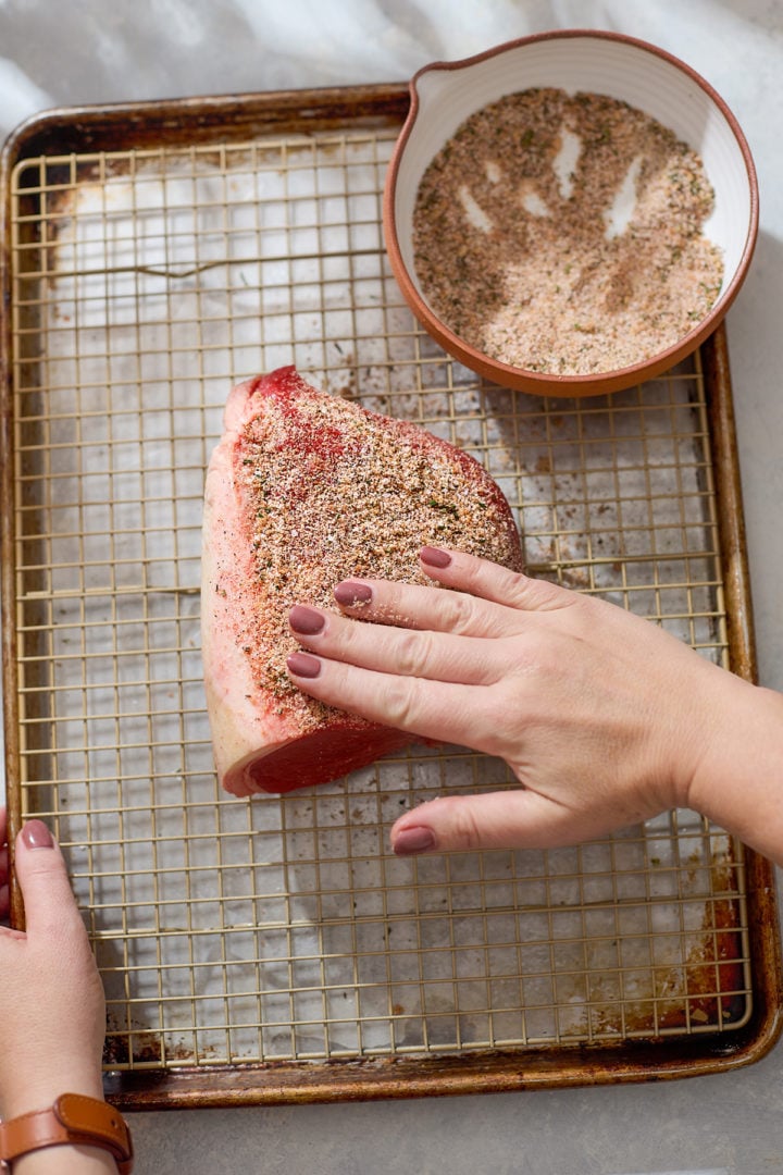 Hands press the spice blend into the surface of the eye of round roast to ensure even coverage. The roast rests on a rack-lined baking sheet, and the spice mixture bowl sits nearby with some of the rub missing, showing it has been used.