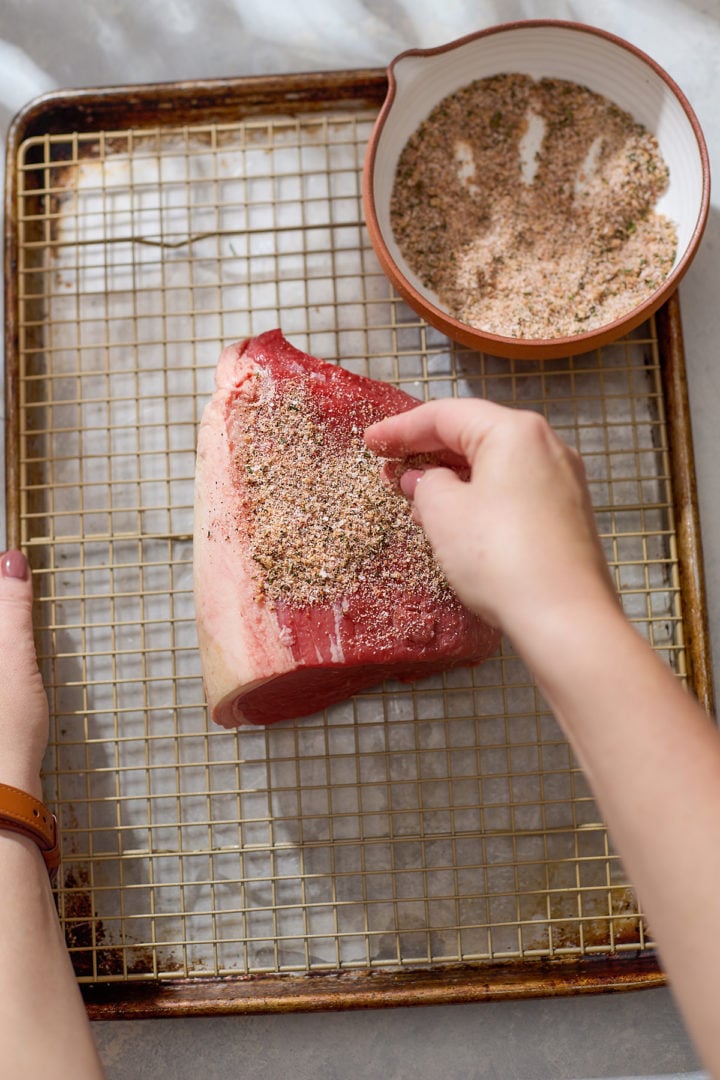 A person’s hands sprinkle the seasoning mixture over the raw beef roast placed on a wire rack set inside a baking sheet. The seasoning bowl is next to the roast, and the surface of the meat is starting to be evenly coated.