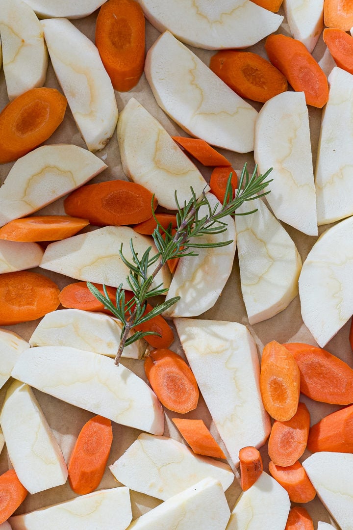 Sliced raw parsnips and carrots arranged on a sheet pan with a sprig of fresh rosemary before roasting.