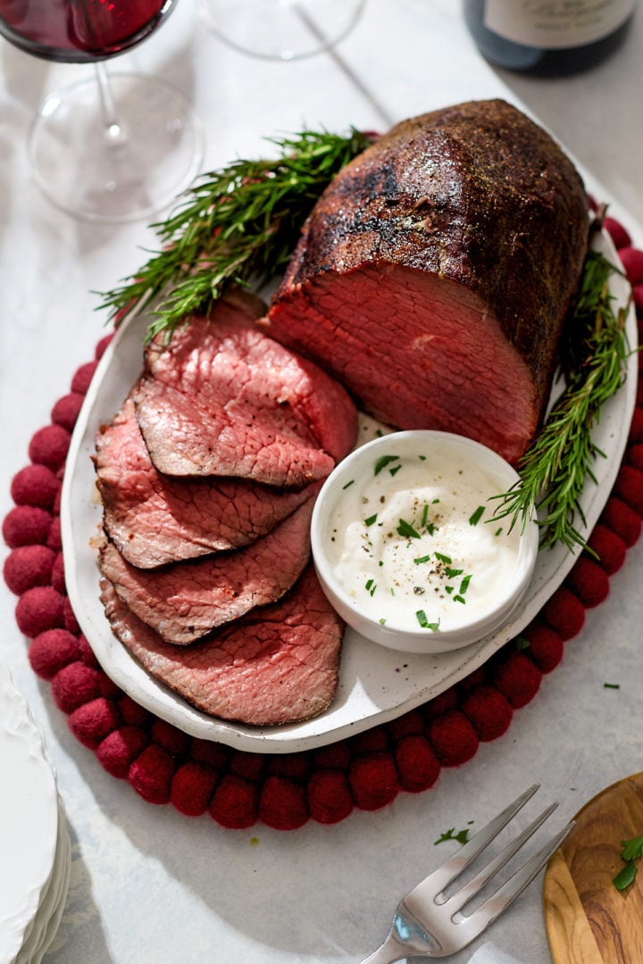 A sliced eye of round roast beef is arranged on an oval white platter with fresh rosemary sprigs and a small bowl of creamy horseradish sauce. The slices reveal a tender, medium-rare pink center. Glasses of red wine sit in the background, creating a festive dinner setting.