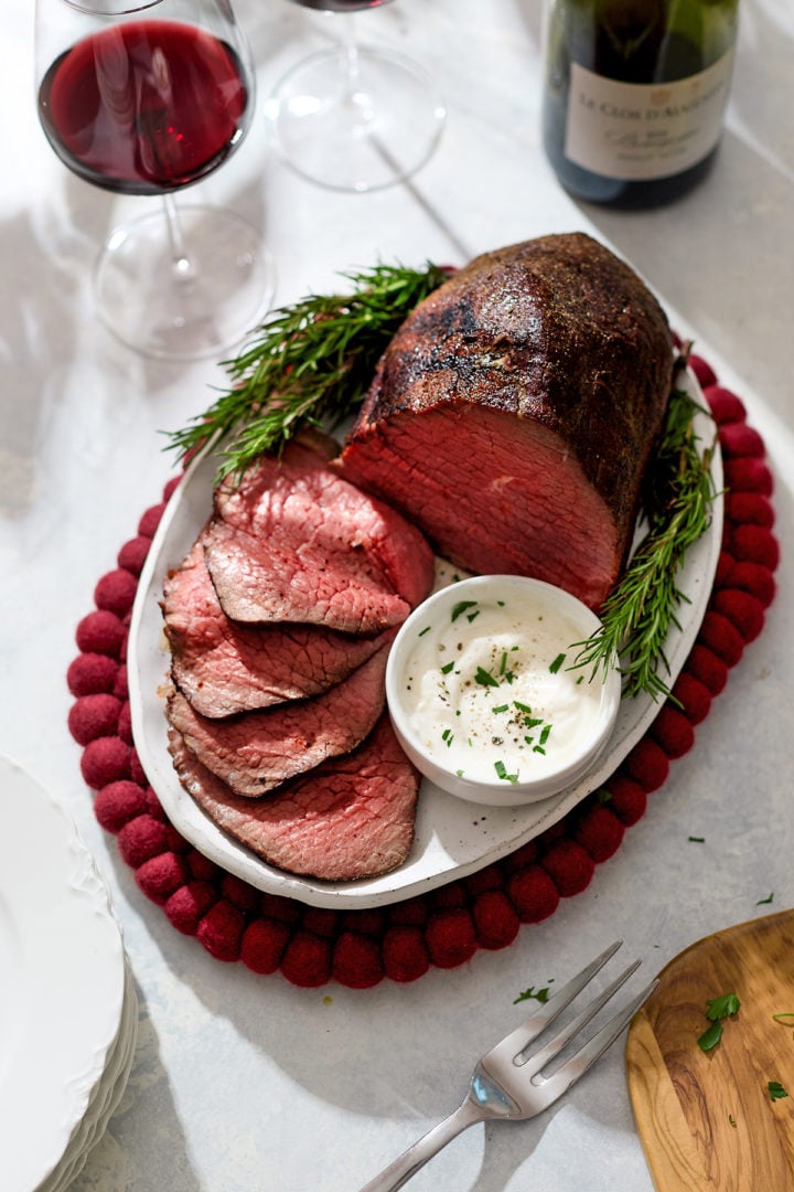 A platter of rosemary roast beef eye of round sits on a red felt trivet, surrounded by fresh rosemary sprigs. The roast is partially sliced, revealing a pink, medium-rare center with a browned crust. Next to the slices is a small bowl of creamy horseradish sauce sprinkled with herbs. Two glasses of red wine and a wine bottle are in the background, creating an elegant holiday dinner setting.