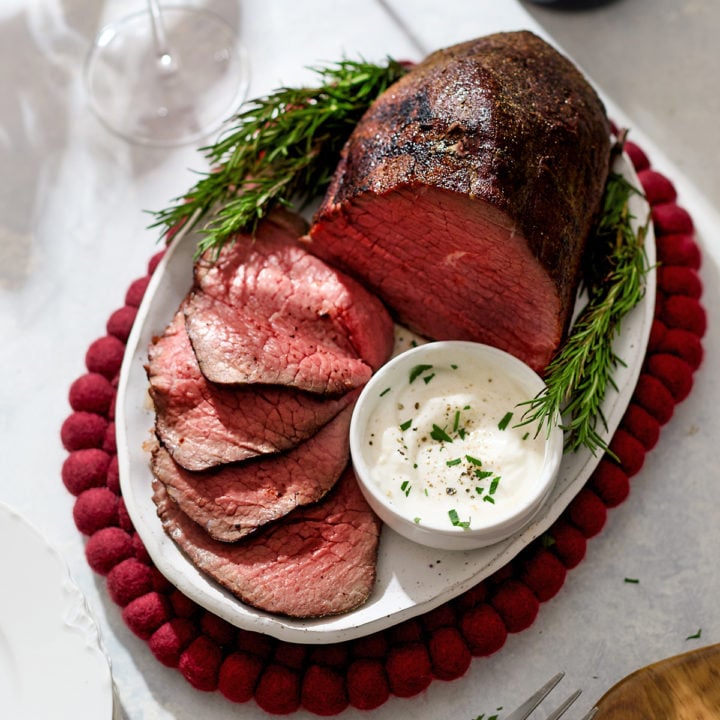 A platter of rosemary roast beef eye of round sits on a red felt trivet, surrounded by fresh rosemary sprigs. The roast is partially sliced, revealing a pink, medium-rare center with a browned crust. Next to the slices is a small bowl of creamy horseradish sauce sprinkled with herbs. Two glasses of red wine and a wine bottle are in the background, creating an elegant holiday dinner setting.