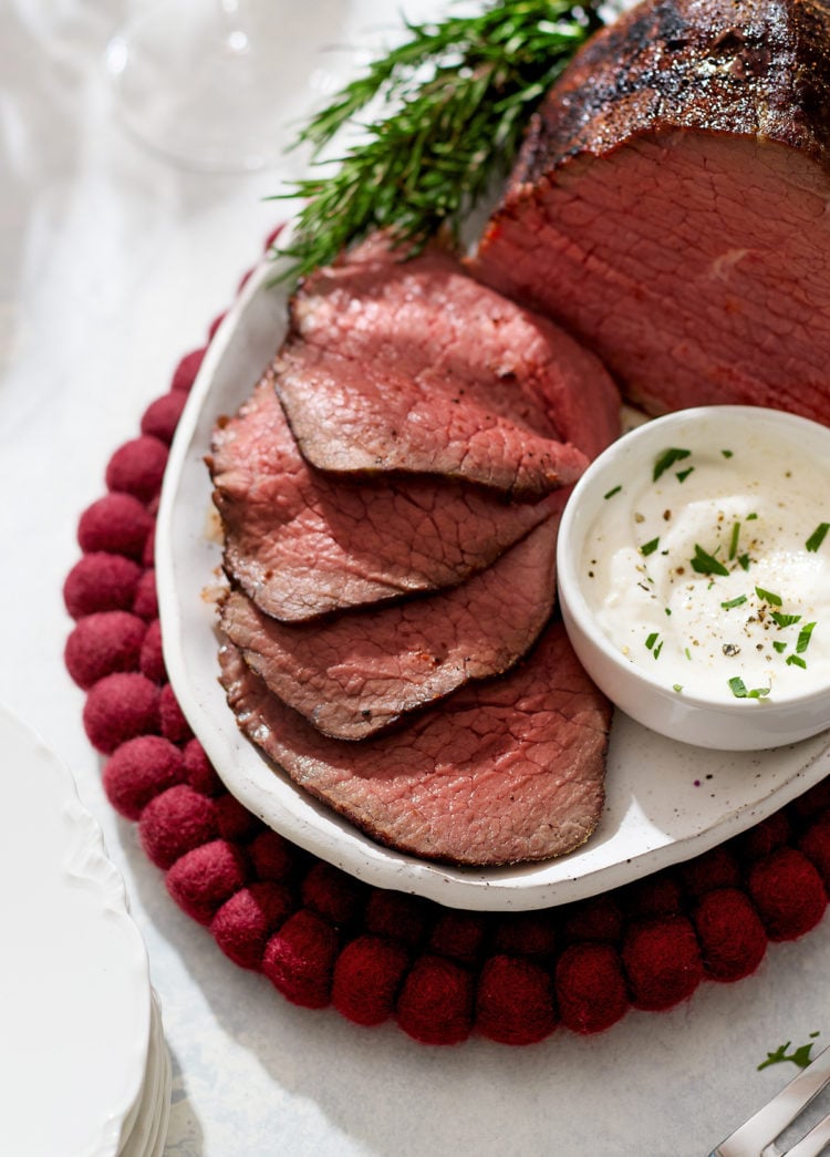 A close-up of thinly sliced roast beef eye of round arranged neatly on a white platter beside a small bowl of horseradish sauce. The meat’s interior is tender and rosy, contrasting with the dark, seared crust. Fresh rosemary sprigs frame the platter, and a red felt trivet peeks from underneath, adding a festive touch.