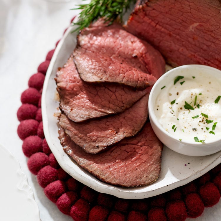 A close-up of thinly sliced roast beef eye of round arranged neatly on a white platter beside a small bowl of horseradish sauce. The meat’s interior is tender and rosy, contrasting with the dark, seared crust. Fresh rosemary sprigs frame the platter, and a red felt trivet peeks from underneath, adding a festive touch.