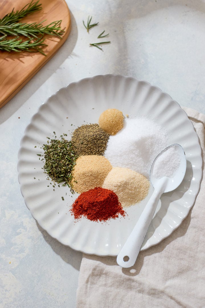 A white fluted plate holds piles of individual seasonings for the dry rub: salt, brown sugar, black pepper, garlic powder, onion powder, paprika, and dried rosemary. A white spoon rests on the plate, and a few rosemary sprigs are visible on a wooden board nearby.