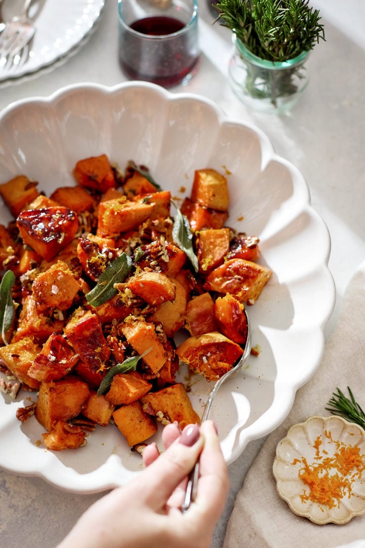 A white scalloped serving bowl filled with roasted candied yams glistening with a maple syrup glaze. The orange-brown cubes are sprinkled with toasted pecans, crisp sage leaves, and orange zest. A hand holding a spoon is serving a portion. In the background sit glasses of red wine, fresh rosemary in a jar, and soft natural sunlight.