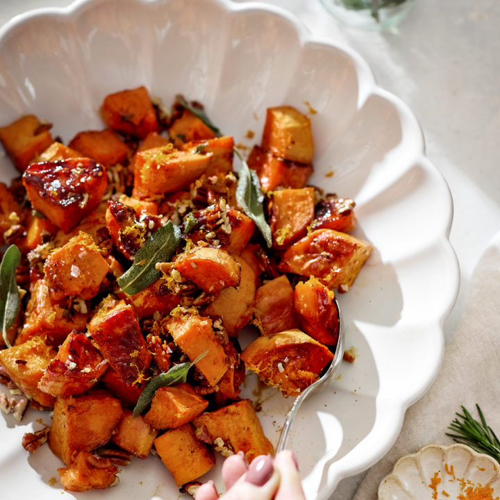A white scalloped serving bowl filled with roasted candied yams glistening with a maple syrup glaze. The orange-brown cubes are sprinkled with toasted pecans, crisp sage leaves, and orange zest. A hand holding a spoon is serving a portion. In the background sit glasses of red wine, fresh rosemary in a jar, and soft natural sunlight.