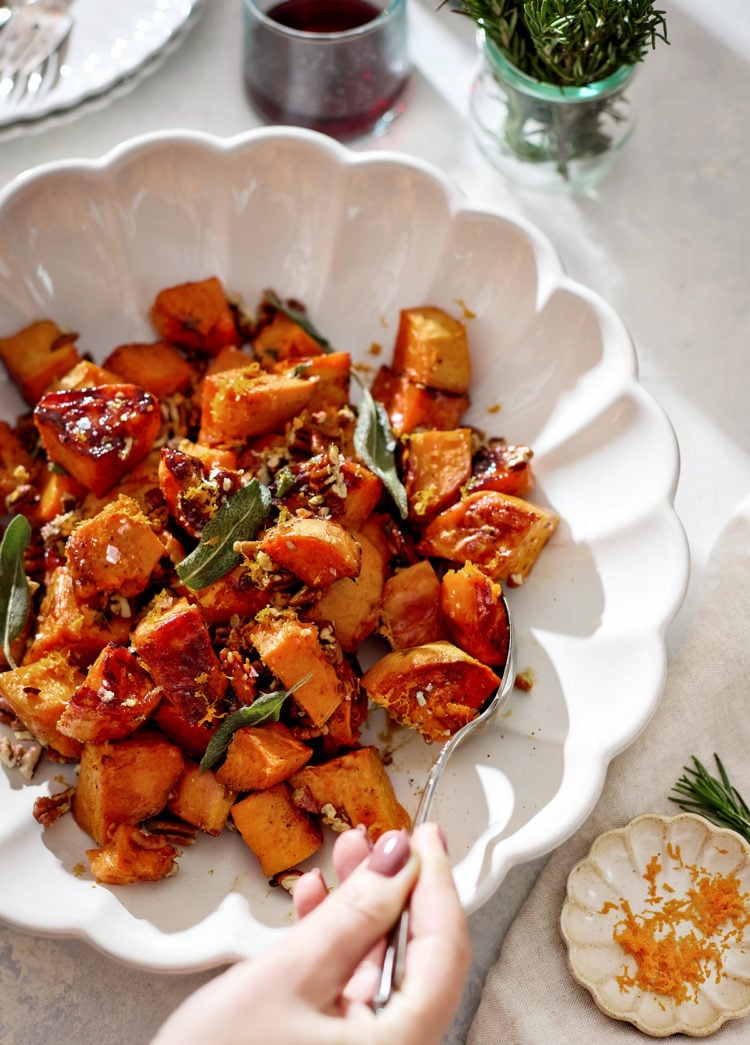 A white scalloped serving bowl filled with roasted candied yams glistening with a maple syrup glaze. The orange-brown cubes are sprinkled with toasted pecans, crisp sage leaves, and orange zest. A hand holding a spoon is serving a portion. In the background sit glasses of red wine, fresh rosemary in a jar, and soft natural sunlight.