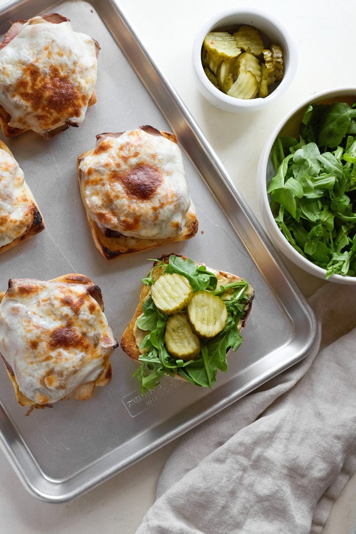 Slices of spicy horseradish pickles are layered onto the arugula on one sandwich half, ready to be topped with the melted-cheese half.
