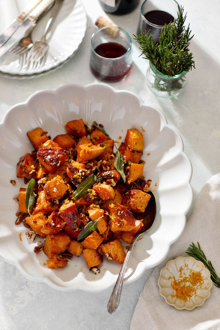 A close-up of baked candied yams tossed with maple syrup, toasted pecans, and fried sage leaves in a large white bowl. The caramelized edges of the yams glisten under natural light, with flecks of sea salt and orange zest scattered across the top.