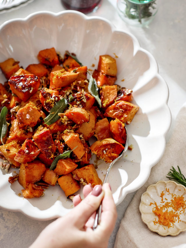 A white scalloped serving bowl filled with roasted candied yams glistening with a maple syrup glaze. The orange-brown cubes are sprinkled with toasted pecans, crisp sage leaves, and orange zest. A hand holding a spoon is serving a portion. In the background sit glasses of red wine, fresh rosemary in a jar, and soft natural sunlight.