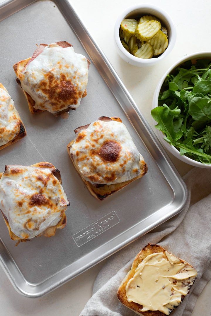 Open-faced sandwiches on a sheet pan after broiling, with the provolone melted and browned on top. Nearby are bowls of arugula and sliced pickles.
