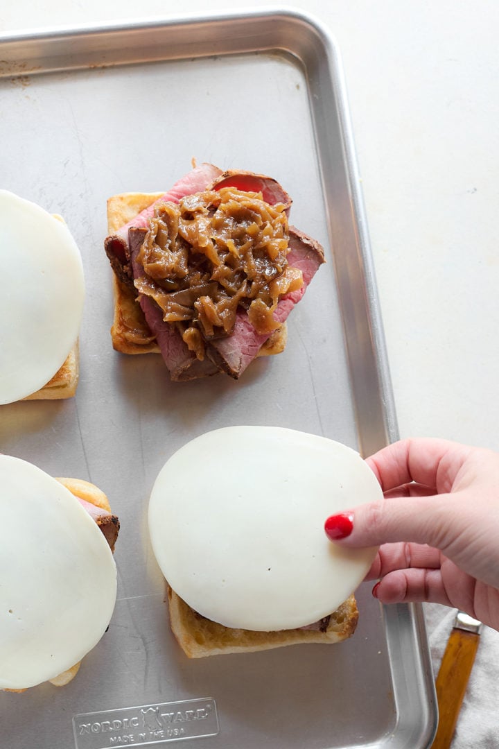 A hand places a slice of provolone cheese on top of the roast beef and caramelized onions on an open-faced sandwich. Other sandwiches already have cheese added.
