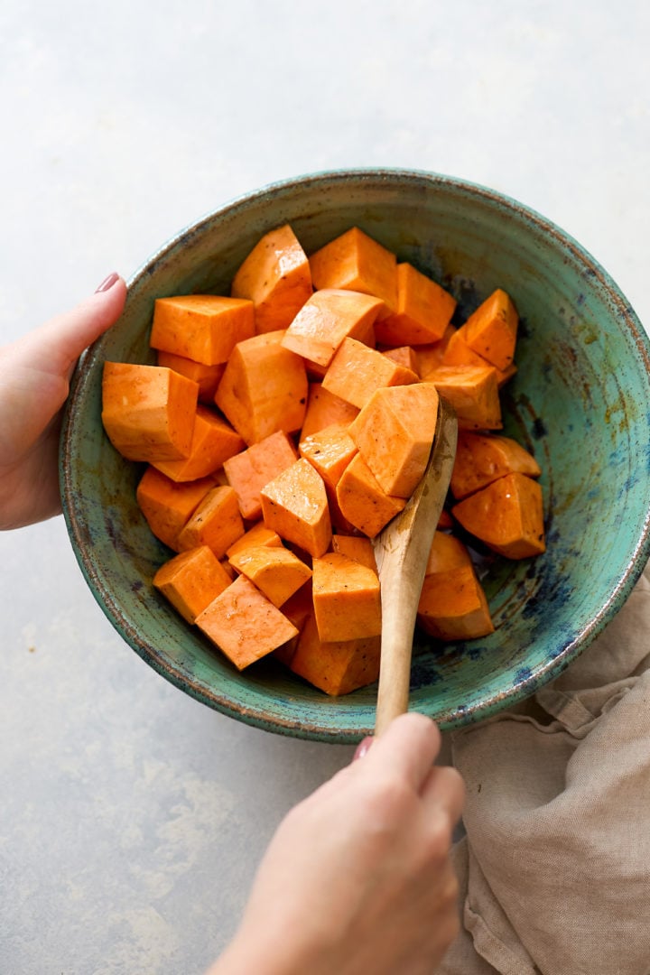 Hands are tossing large chunks of peeled yams in a teal-green mixing bowl with a wooden spoon. The yams are coated in a shiny maple brown-butter mixture made with orange zest and warm spices, but there are no pecans or sage on them yet. The bowl sits on a light countertop with a beige linen underneath, showing the step just before the yams are spread on the pan to roast.