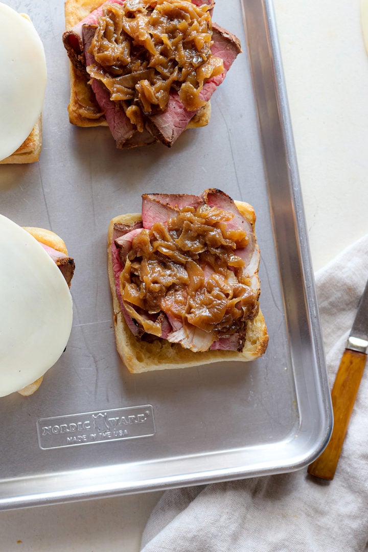 Open-faced ciabatta rolls on a sheet pan topped with slices of roast beef. One sandwich has a generous spoonful of caramelized onions layered on top.
