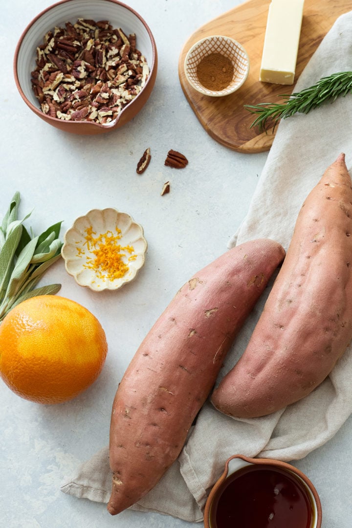 All ingredients arranged on a light countertop: whole sweet potatoes, an orange, a small dish of zest, a bowl of chopped pecans with bits of sage, a stick of butter on a wooden board, a sprig of rosemary, a small bowl of maple syrup, and a bowl of ground cinnamon. A beige linen napkin softens the scene.
