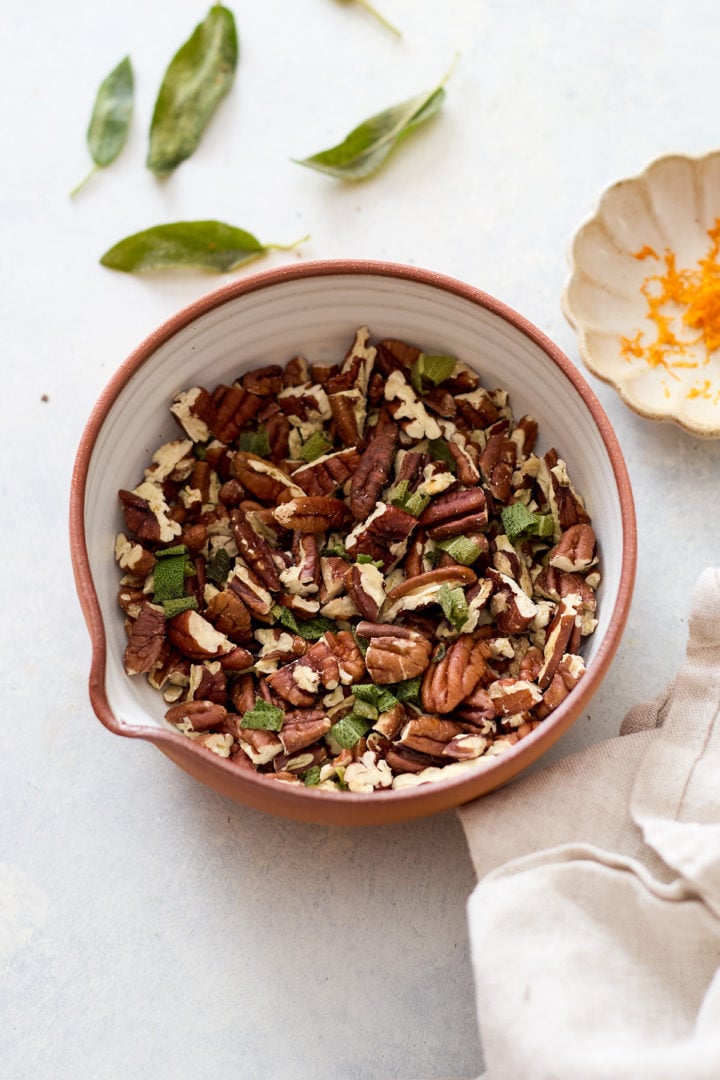 Chopped pecans and finely sliced sage leaves fill a ceramic bowl. Around it, loose sage leaves and a small dish of bright orange zest rest on the table, with a neutral fabric napkin beside them.