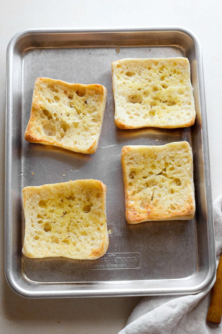 Four ciabatta roll halves sit cut-side up on a baking sheet, lightly toasted and glistening with oil or melted butter.
