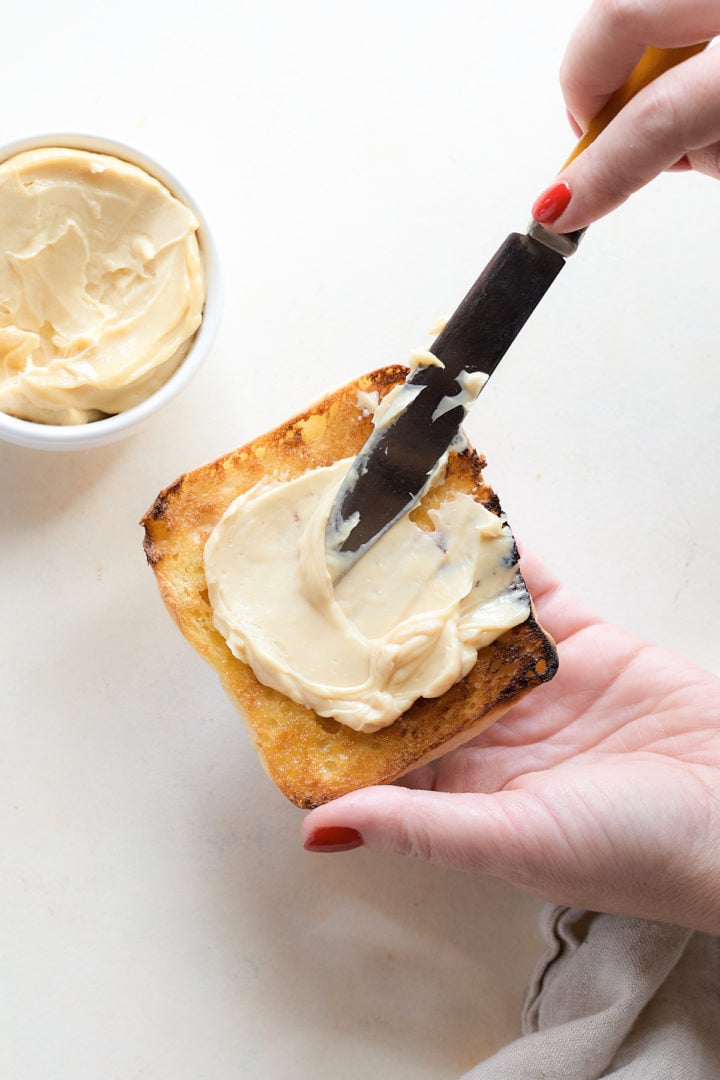 A hand spreads creamy garlic aioli onto the toasted cut side of a ciabatta roll, with the bowl of aioli next to it.
