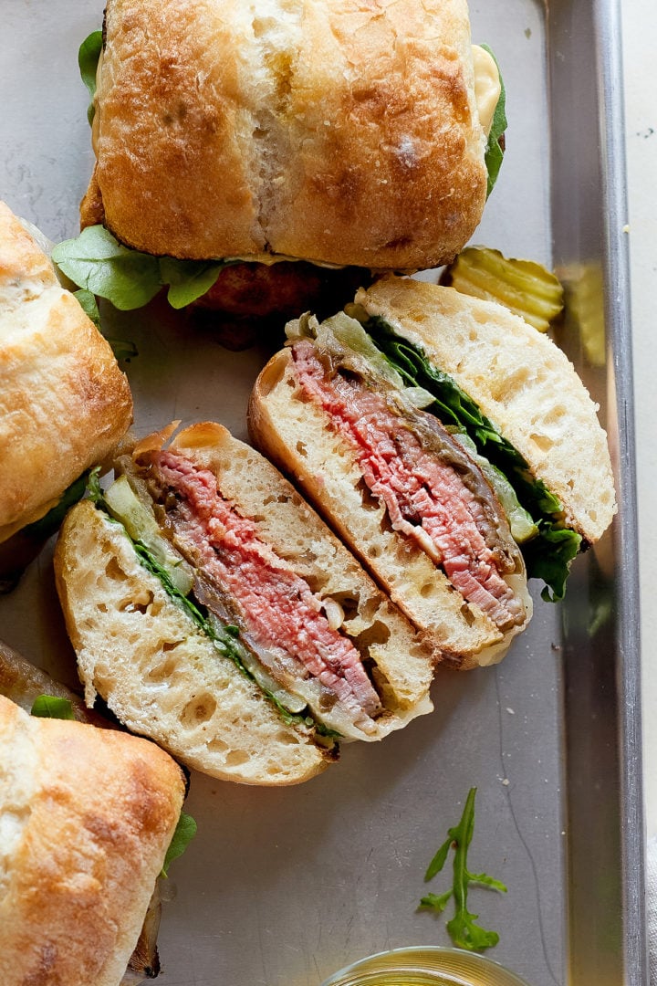 A hot roast beef sandwich is sliced in half to show the inside layers: juicy roast beef, melted provolone, caramelized onions, arugula, and pickles inside a toasted ciabatta roll. Other whole sandwiches sit nearby on the sheet pan.
