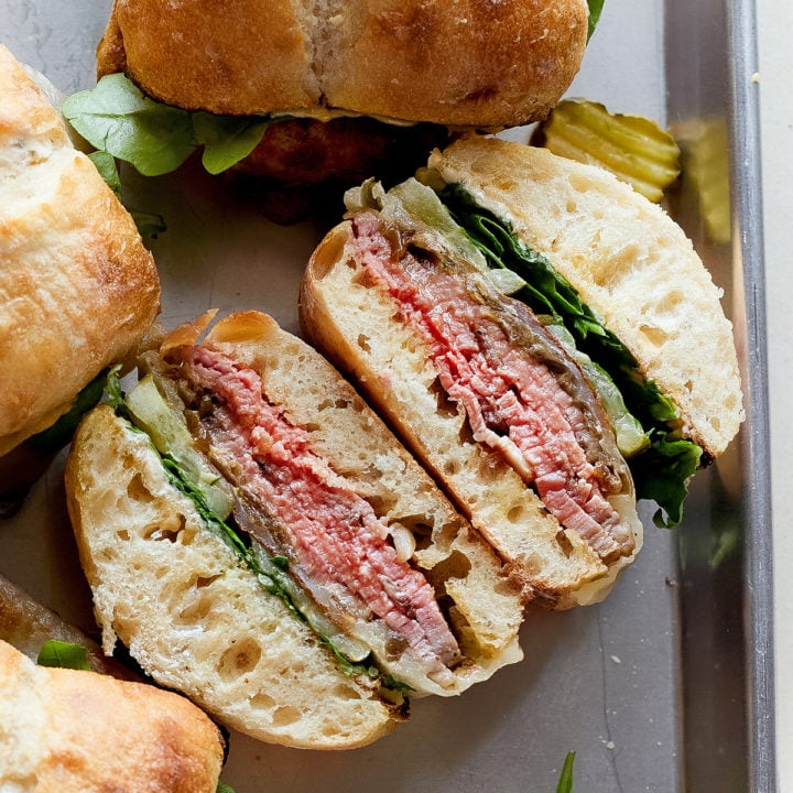 A hot roast beef sandwich is sliced in half to show the inside layers: juicy roast beef, melted provolone, caramelized onions, arugula, and pickles inside a toasted ciabatta roll. Other whole sandwiches sit nearby on the sheet pan.