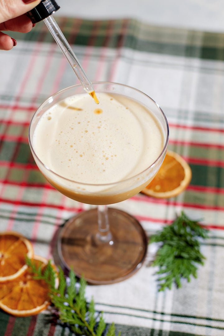 A dropper releasing aromatic bitters into a foamy gingerbread whiskey sour cocktail in a coupe glass. The drink is set on a holiday plaid tablecloth with dried orange slices and evergreen sprigs for decoration.