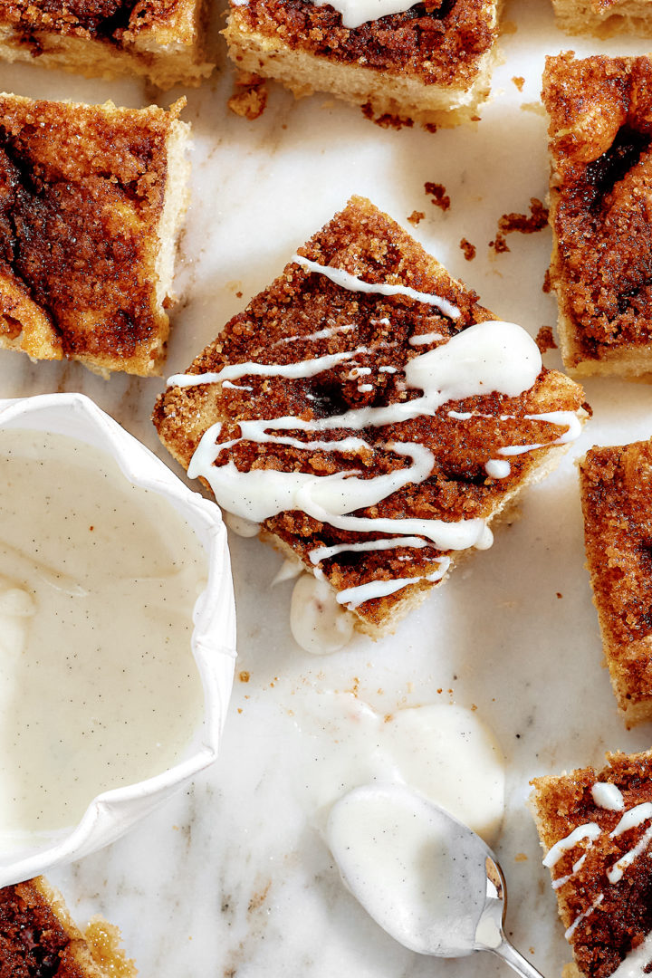 Sliced cinnamon sugar focaccia arranged on a marble surface with white glaze drizzled over the top. A small white bowl of glaze and a spoon sit nearby.