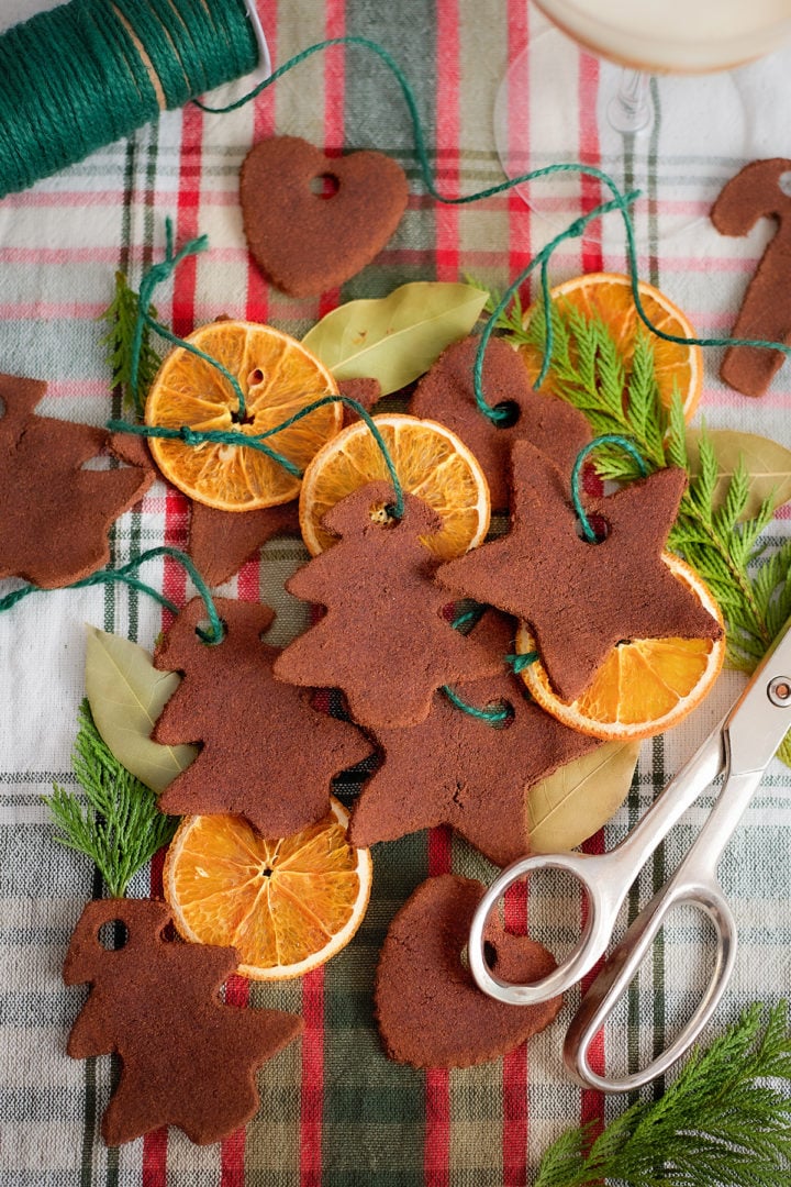 Assorted dried orange slices and cinnamon dough ornaments are arranged on a red and green plaid cloth. The ornaments are cut into shapes like trees, stars, hearts, and candy canes. Each ornament has a hole punched near the top and is tied with green string. Bay leaves, evergreen sprigs, and a pair of silver scissors are also included in the scene.