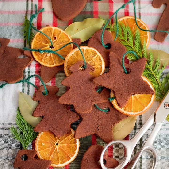 Assorted dried orange slices and cinnamon dough ornaments are arranged on a red and green plaid cloth. The ornaments are cut into shapes like trees, stars, hearts, and candy canes. Each ornament has a hole punched near the top and is tied with green string. Bay leaves, evergreen sprigs, and a pair of silver scissors are also included in the scene.