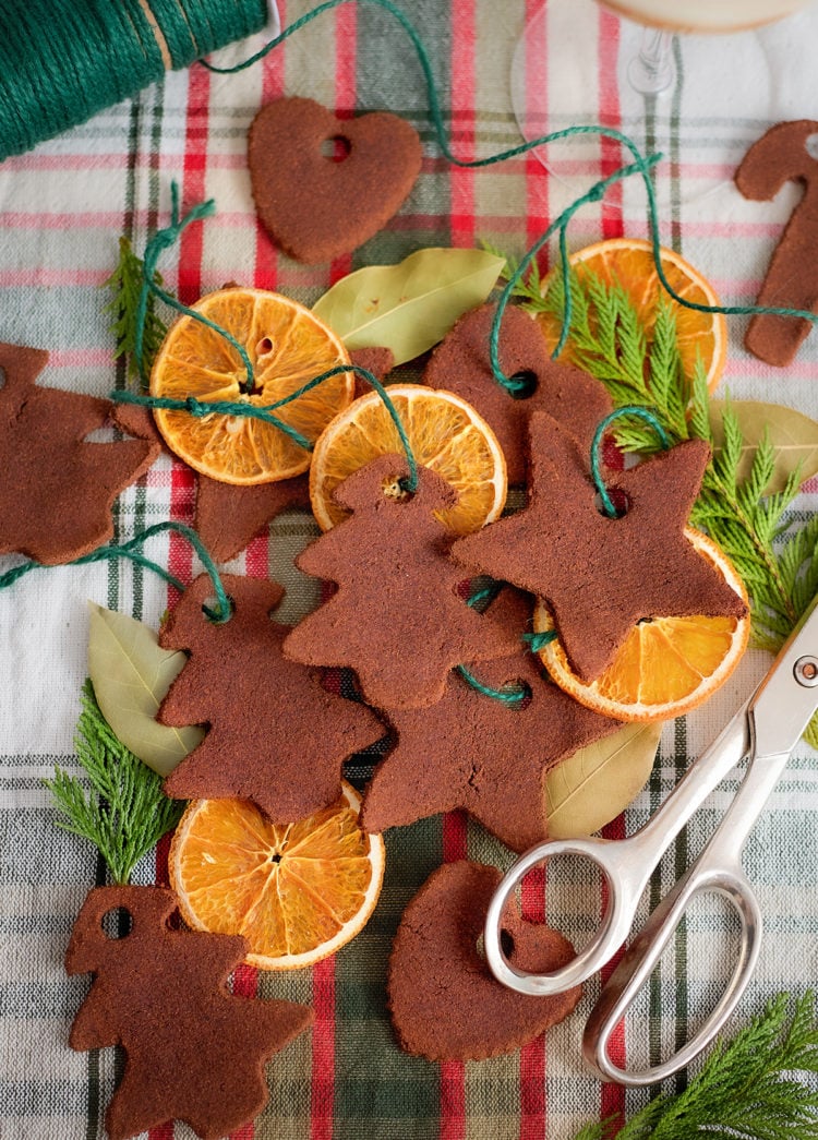 Assorted dried orange slices and cinnamon dough ornaments are arranged on a red and green plaid cloth. The ornaments are cut into shapes like trees, stars, hearts, and candy canes. Each ornament has a hole punched near the top and is tied with green string. Bay leaves, evergreen sprigs, and a pair of silver scissors are also included in the scene.