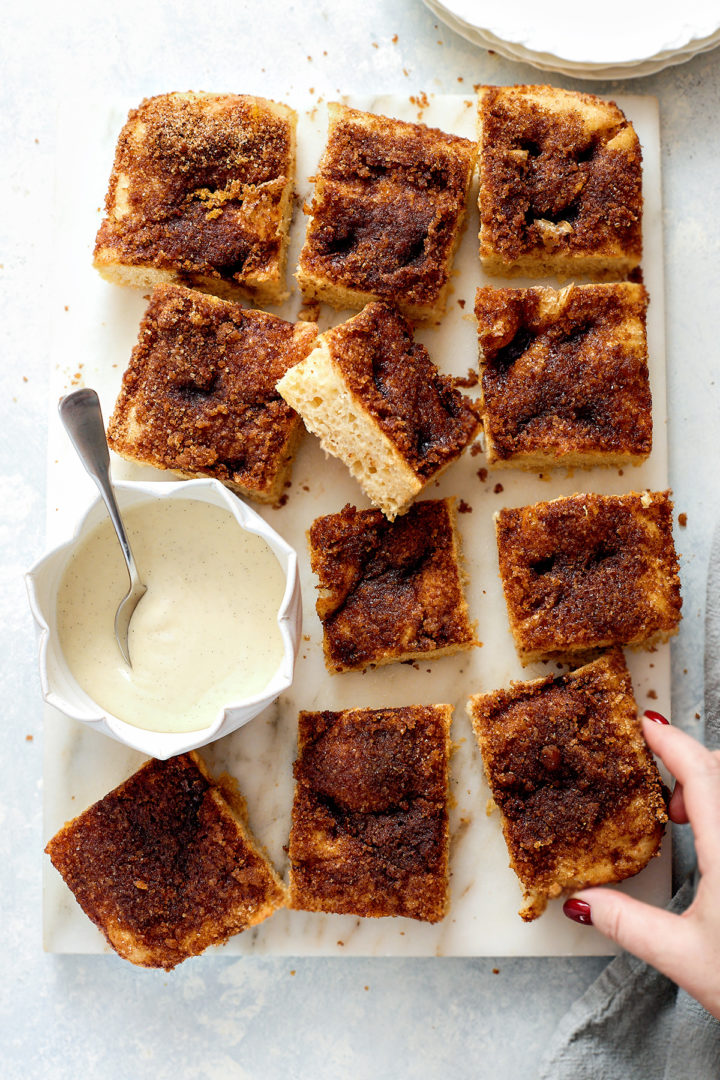 Overhead view of cinnamon sugar focaccia cut into squares and arranged on a white marble board. Some pieces are stacked slightly or leaning. A small white bowl of vanilla glaze with a spoon is placed to the side, and a hand is reaching in to grab a slice.