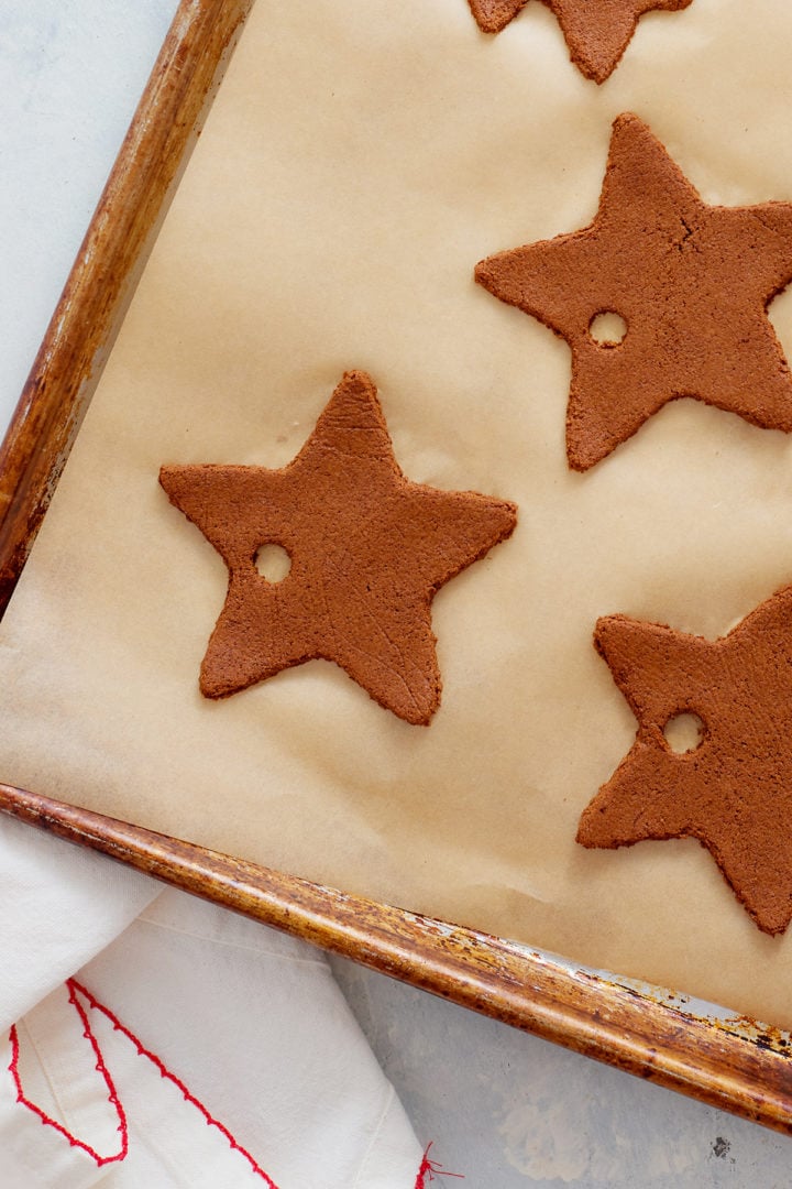 Close-up view of star-shaped cinnamon ornaments arranged on a parchment-lined baking sheet. The ornaments have a rough, matte texture and a small circular hole near the top of each one. A white cloth with red stitching is visible in the bottom left corner.