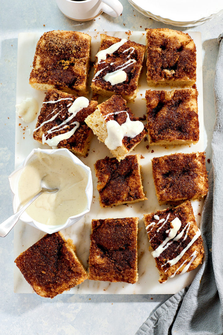 Overhead view of cinnamon focaccia squares with glaze drizzled on some pieces. A white coffee cup appears at the top of the frame.
