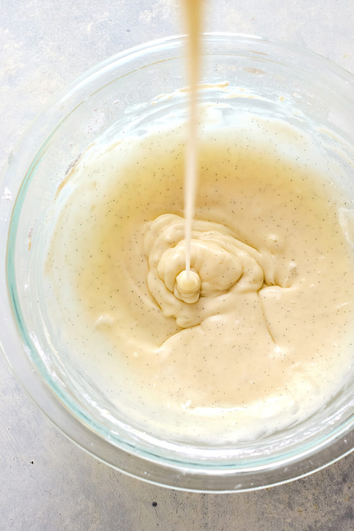 Close-up overhead shot of creamy glaze being poured into a glass bowl, forming thick ribbons as it lands on the surface of the glaze.