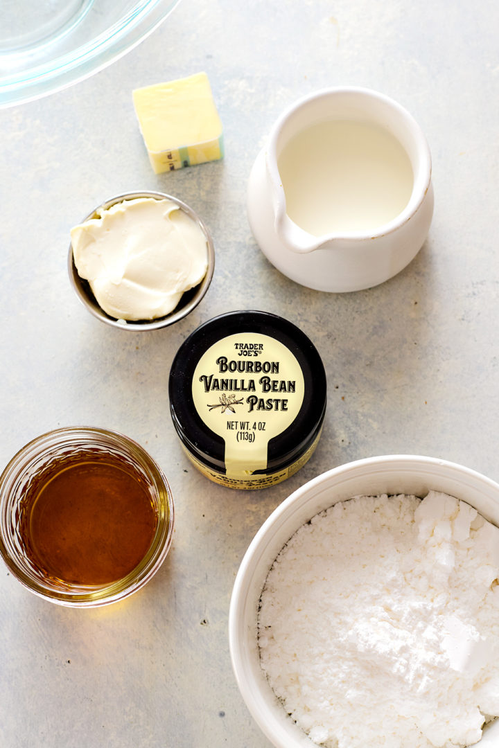 Overhead view of glaze ingredients arranged on a light surface: a bowl of powdered sugar, a small bowl of cream cheese, a small bowl of melted butter, a container of vanilla bean paste, a measuring cup of milk, and a small dish of bourbon.