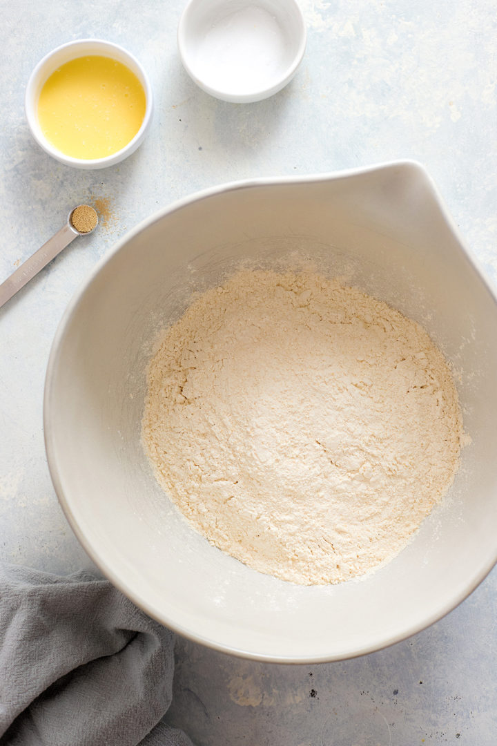 Overhead view of a mixing bowl with flour, surrounded by small bowls of active dry yeast, fine salt, and melted butter, showing ingredients for sweet focaccia dough.
