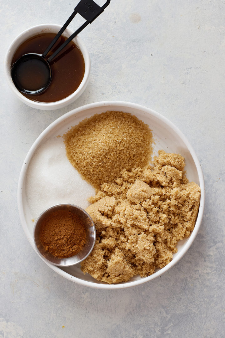 A white plate filled with granulated sugar, light brown sugar, turbinado sugar, and ground cinnamon, alongside a bowl of browned butter with black measuring spoons.